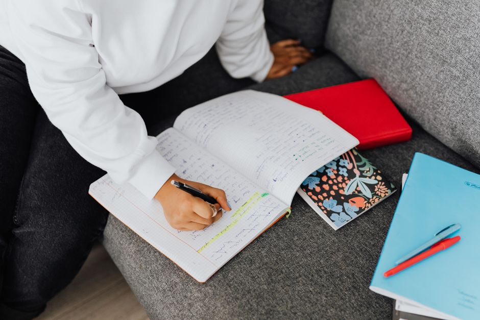 A woman in a white sweater writes notes on a notebook while studying on a sofa.