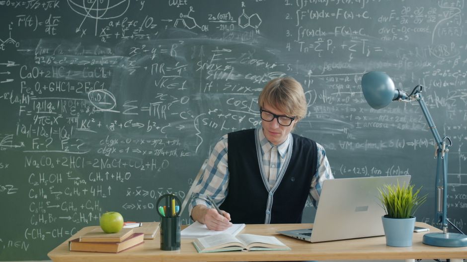 Teacher in classroom writing on notebook, surrounded by books, laptop, and chalkboard.